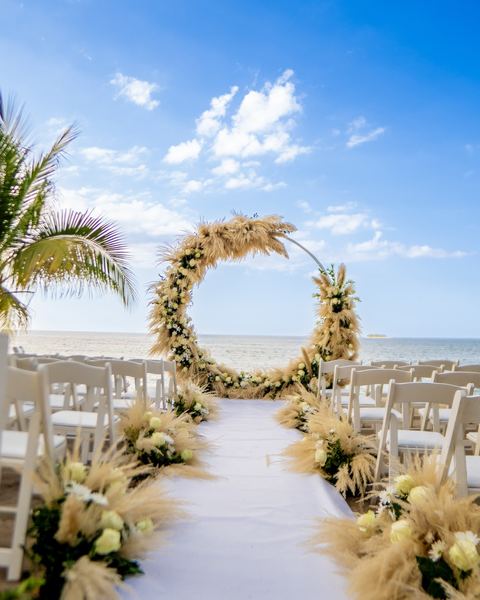 Allée de cérémonie sur la plage, devant une arche ronde de fleurs et de pampa